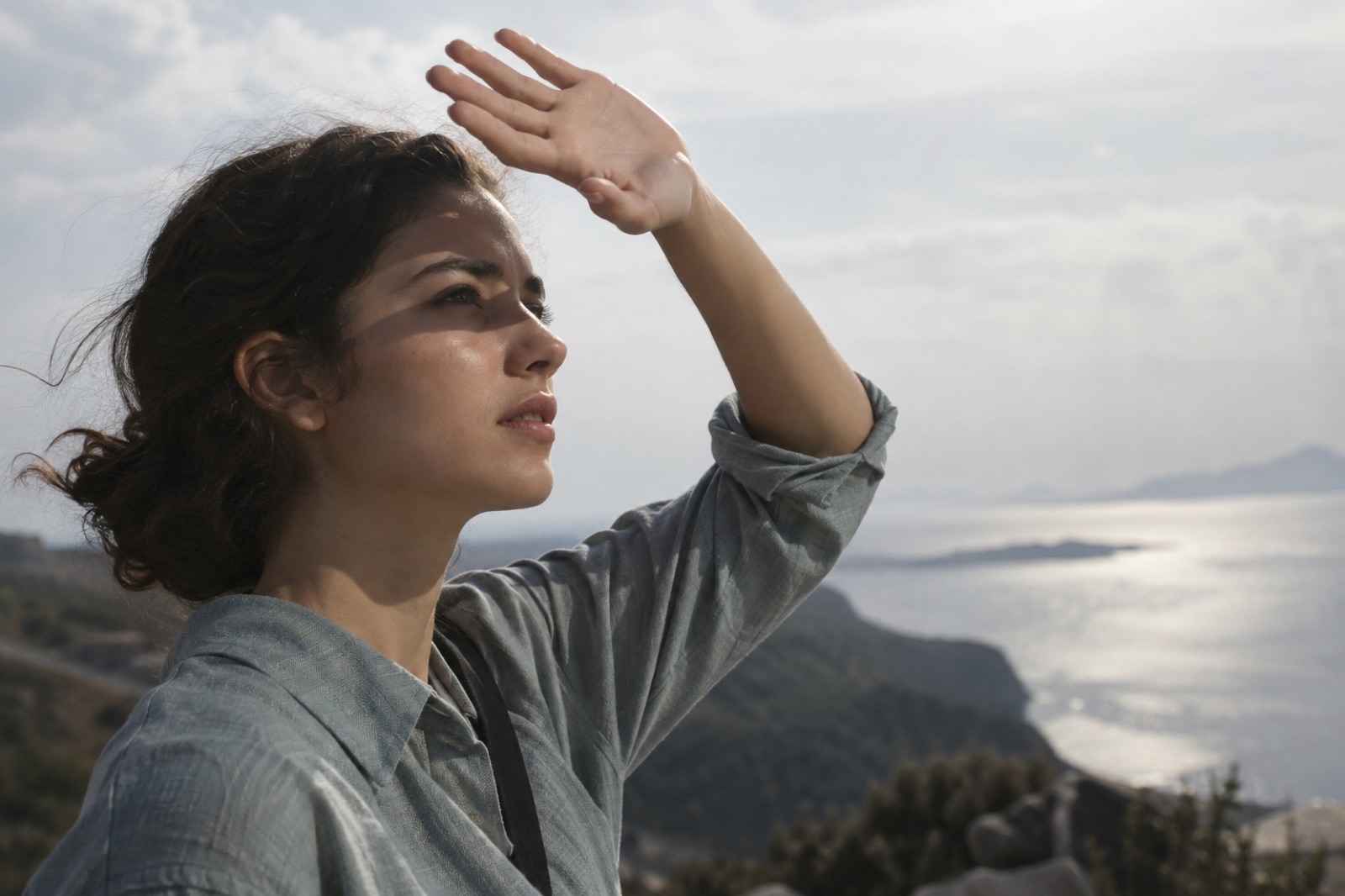 Side view of a woman on a hill overlooking the sea, shading her eyes with one hand as she looks into the distance