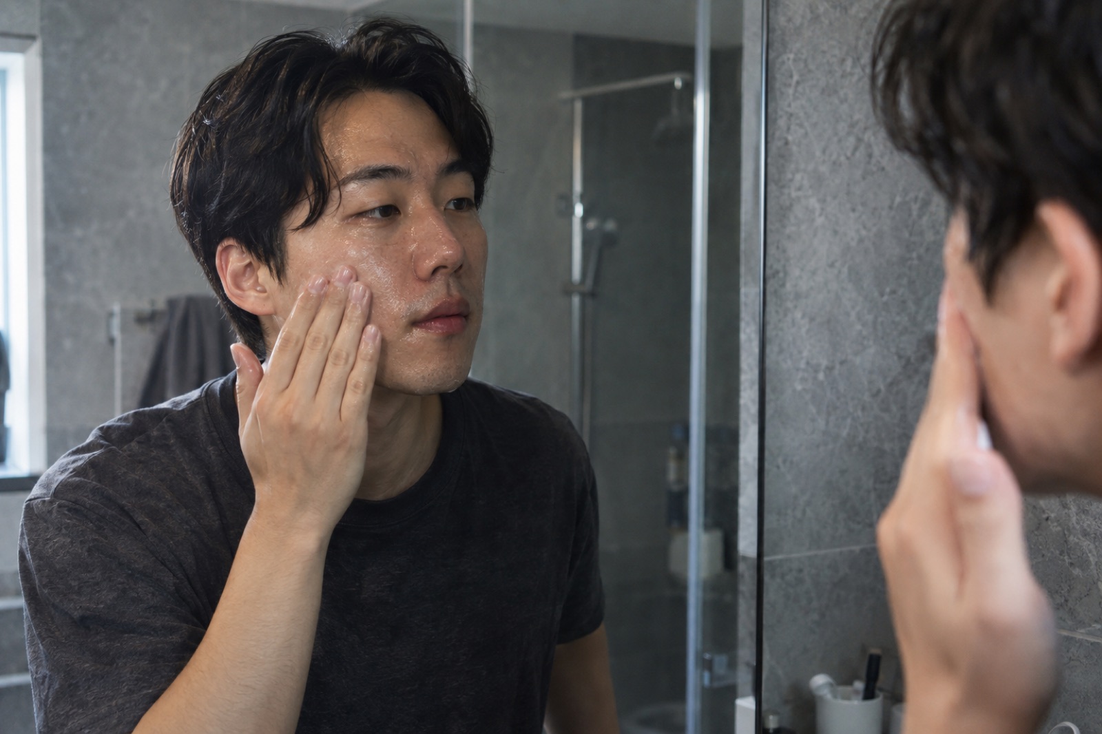 A man in a black T-shirt in front of a bathroom mirror with gray tile, touching his damp cheek with his fingertips