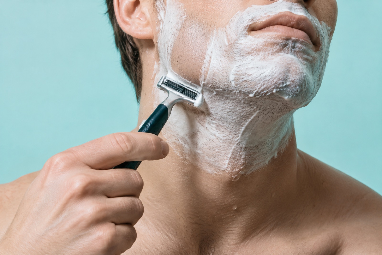Một người đàn ông trước phông nền xanh nhạt, cằm và cổ phủ bọt trắng, đang đưa dao cạo dọc phía dưới đường viền hàm